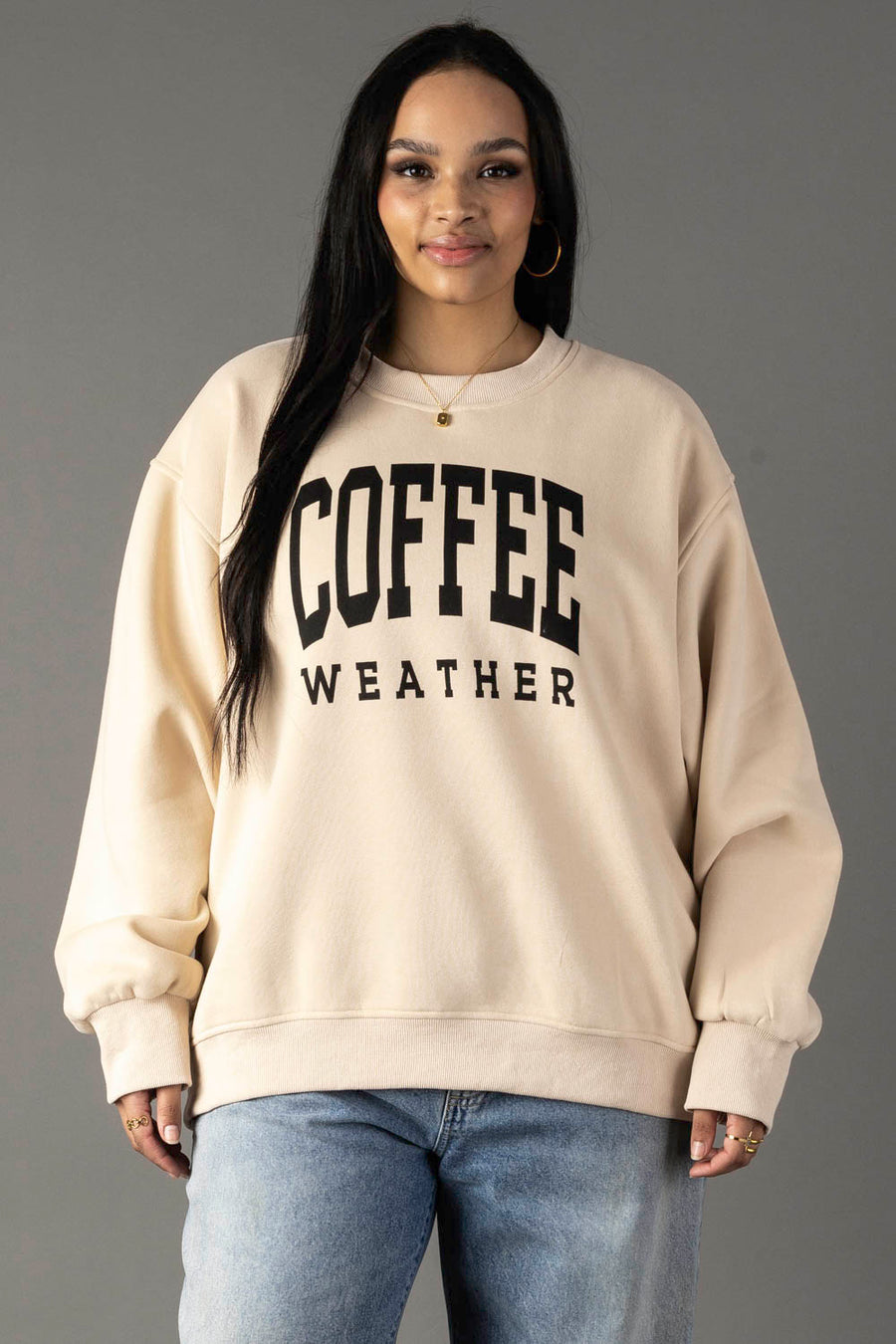 A woman with long dark hair wears a Tan Coffee Weather Sweatshirt from Panache Apparel Collection and light blue jeans, standing against a plain gray background.