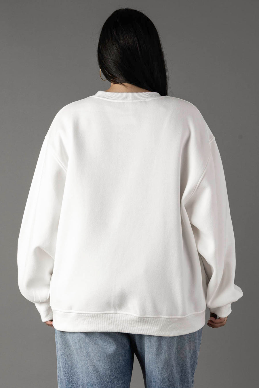 A person with long dark hair is seen from behind against a gray background, wearing the Panache Apparel Collection’s oversized White Coffee Weather Sweatshirt paired with blue jeans.