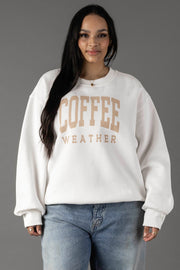 A woman with long dark hair wears the Panache Apparel Collection White Coffee Weather Sweatshirt, featuring beige lettering. She pairs the cozy fleece pullover with light blue jeans and stands against a plain gray background, looking relaxed.