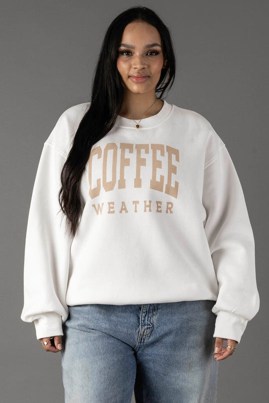 A woman with long dark hair wears the Panache Apparel Collection White Coffee Weather Sweatshirt, featuring beige lettering. She pairs the cozy fleece pullover with light blue jeans and stands against a plain gray background, looking relaxed.