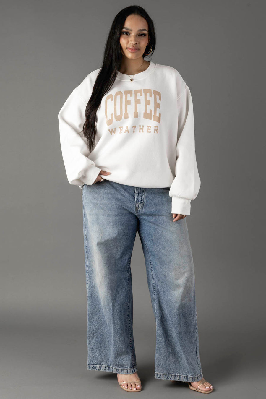 A woman with long dark hair smiles gently at the camera, wearing a Panache Apparel Collection White Coffee Weather Sweatshirt against a gray background, paired with loose blue jeans and beige sandals.
