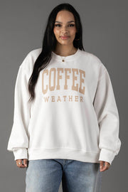 A woman with long dark hair wears the Panache Apparel Collection White Coffee Weather Sweatshirt with light tan lettering, paired with blue jeans, standing against a plain gray background.