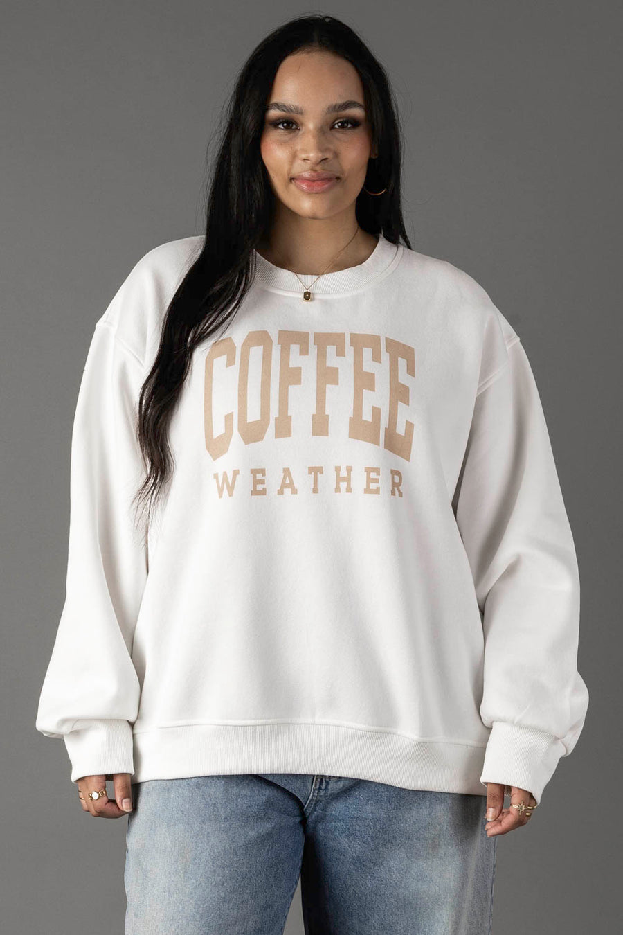 A woman with long dark hair wears the Panache Apparel Collection White Coffee Weather Sweatshirt with light tan lettering, paired with blue jeans, standing against a plain gray background.