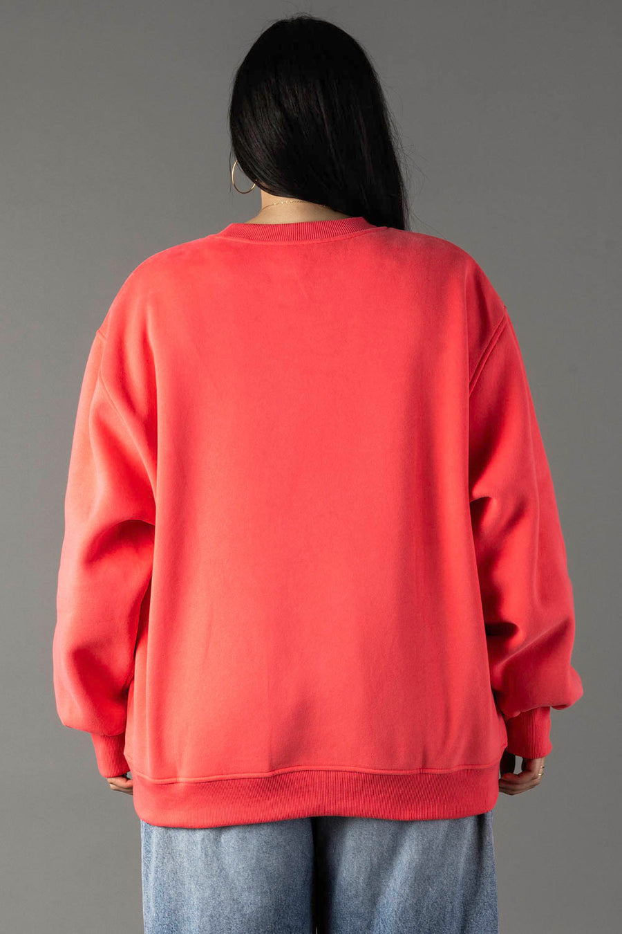 A person with long dark hair wears the Coral Day Drinking Weather Sweatshirt by Panache Apparel Collection and light blue jeans, standing with their back to the camera against a plain gray background.