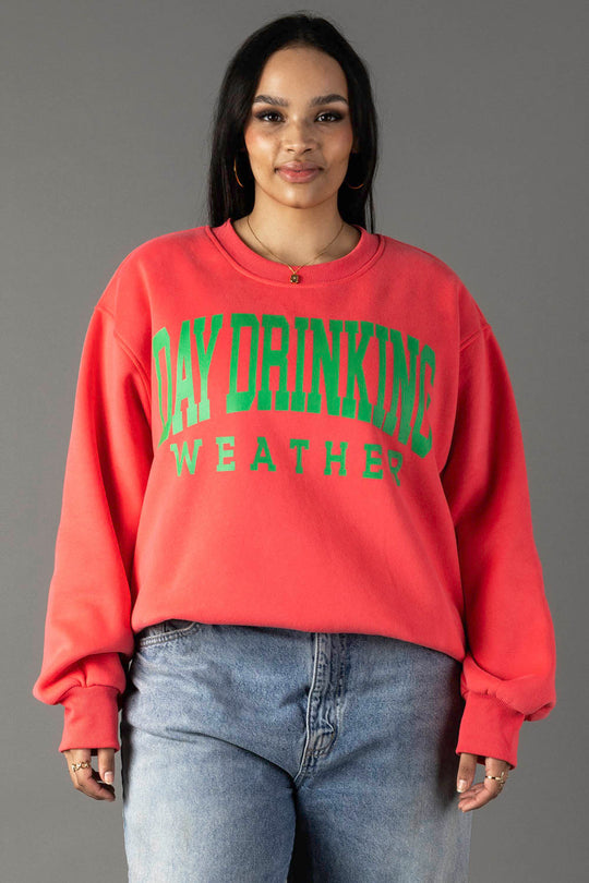 A woman in a Coral Day Drinking Weather Sweatshirt by Panache Apparel Collection, featuring green text, stands against a gray background. She wears blue jeans, gold hoop earrings, and has long dark hair with a slight smile.