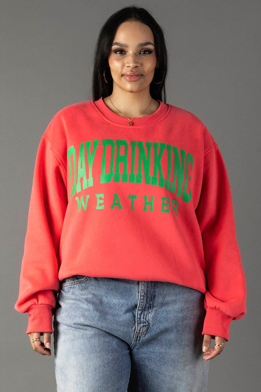 A woman in a Coral Day Drinking Weather Sweatshirt by Panache Apparel Collection, featuring green text, stands against a gray background. She wears blue jeans, gold hoop earrings, and has long dark hair with a slight smile.
