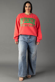A woman smiles at the camera against a gray backdrop, wearing the Panache Apparel Collection Coral Day Drinking Weather Sweatshirt with green lettering, paired with loose blue jeans and open-toed shoes.