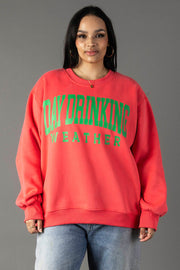 A woman in a Panache Apparel Collection Coral Day Drinking Weather Sweatshirt with green text stands before a gray background. She pairs this comfy fleece crewneck with hoop earrings and blue jeans for laid-back style.