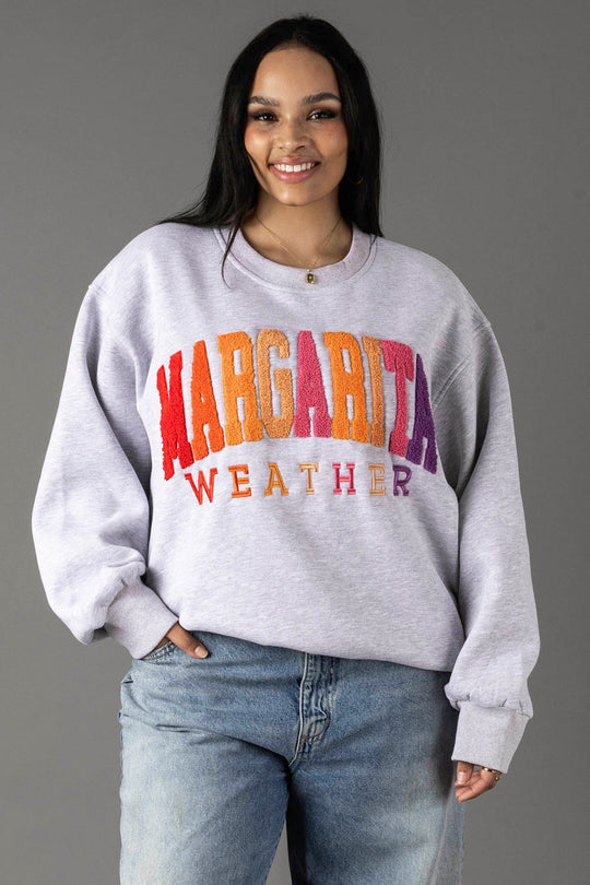 A woman with long dark hair smiles in the Panache Apparel Collection Gray Margarita Weather Sweatshirt with colorful text and blue jeans, standing against a gray background. The soft fleece interior enhances her cozy style.