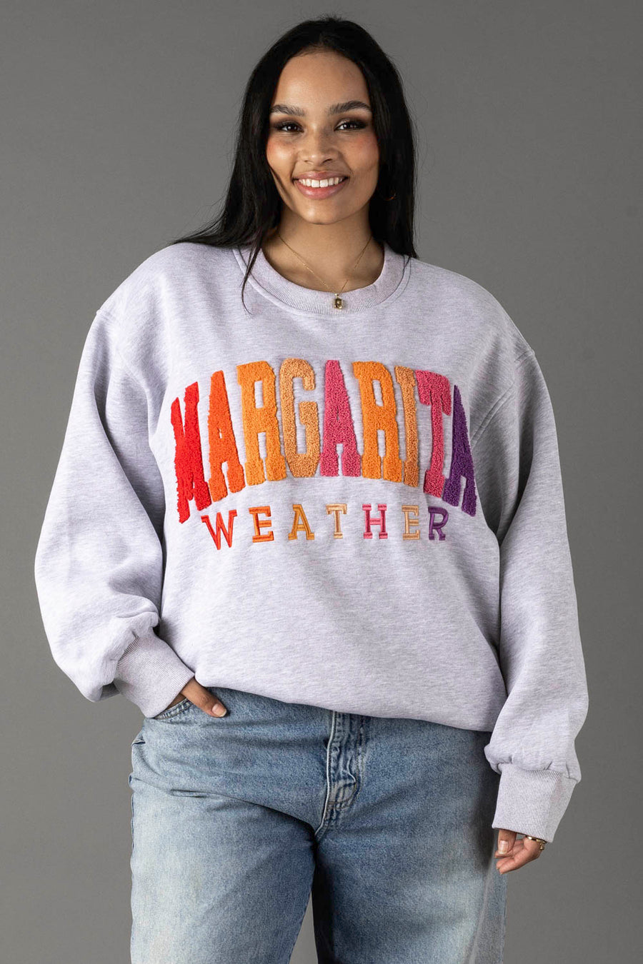A woman with long dark hair smiles in the Panache Apparel Collection Gray Margarita Weather Sweatshirt with colorful text and blue jeans, standing against a gray background. The soft fleece interior enhances her cozy style.