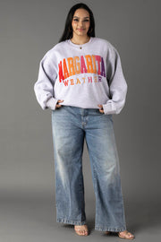 A woman smiles with her hands in her pockets, wearing a Gray Margarita Weather Sweatshirt from Panache Apparel Collection, wide-leg blue jeans, and open-toe heels against a gray background.