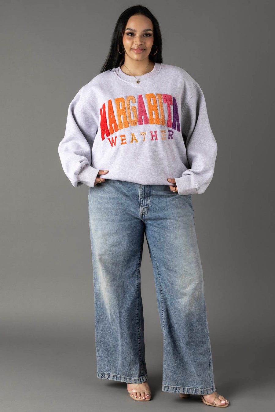 A woman smiles with her hands in her pockets, wearing a Gray Margarita Weather Sweatshirt from Panache Apparel Collection, wide-leg blue jeans, and open-toe heels against a gray background.