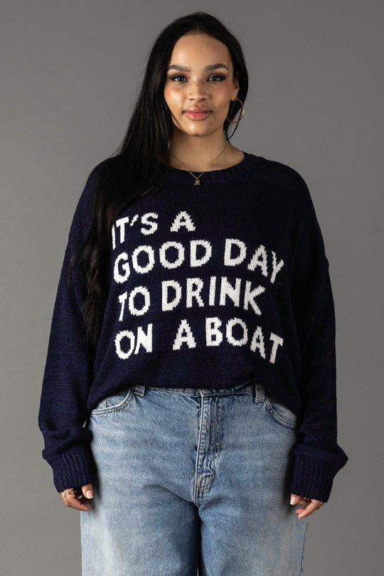 A woman with long dark hair wears the Panache Apparel Collection Drink On A Boat Lightweight Sweater, featuring "It's a good day to drink on a boat" in white on navy blue. She smiles gently at the camera against a gray background.