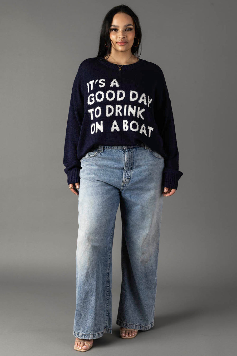 A woman with long dark hair poses against a gray background wearing the Panache Apparel Collection Drink On A Boat Lightweight Sweater in navy and light blue wide-leg jeans, paired with open-toe shoes.