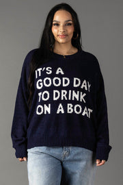 A woman models the Panache Apparel Collection's Drink On A Boat Lightweight Sweater in navy with white "ITS A GOOD DAY TO DRINK ON A BOAT" text, paired with light blue jeans, against a plain gray backdrop.