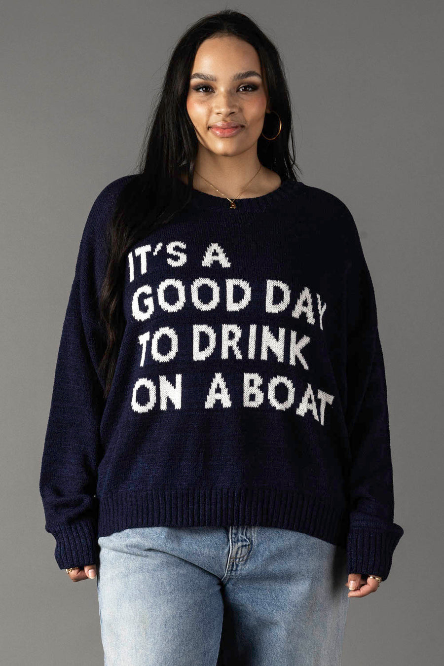 A woman models the Panache Apparel Collection's Drink On A Boat Lightweight Sweater in navy with white "ITS A GOOD DAY TO DRINK ON A BOAT" text, paired with light blue jeans, against a plain gray backdrop.