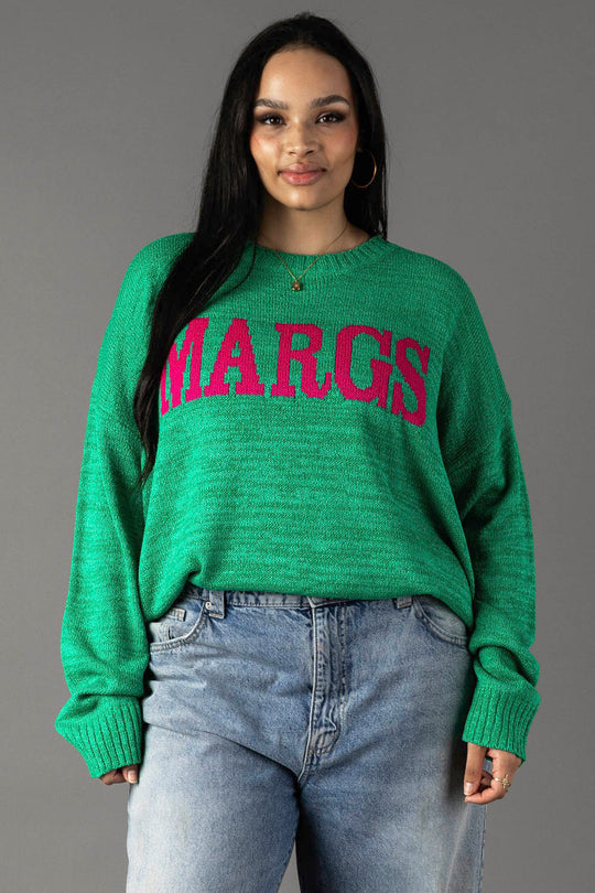 A woman with long dark hair models the Panache Apparel Collection Green Margs Lightweight Sweater, featuring large pink "MARGS" lettering, paired with light blue jeans against a plain gray background. She faces the camera, smiling slightly.