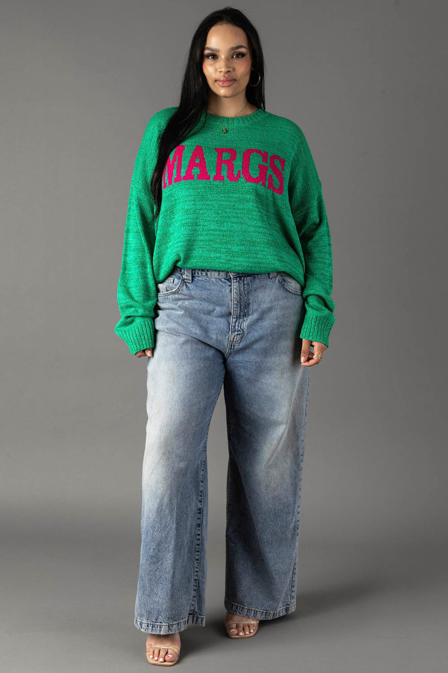 A woman with long dark hair and a neutral expression wears the Panache Apparel Collection Green Margs Lightweight Sweater, featuring pink lettering, paired with loose light blue jeans and open-toed shoes against a gray background.