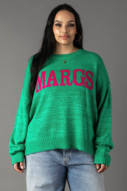 A woman with long dark hair models the Panache Apparel Collection Green Margs Lightweight Sweater, featuring bold pink "MARGS" lettering, paired with light blue jeans against a plain gray background.