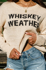 A person wearing the Panache Apparel Collection Tan Whiskey Weather Lightweight Sweater with “WHISKEY WEATHER” text and denim shorts is holding up part of the relaxed-fit sweater, revealing a necklace and rings on their fingers.