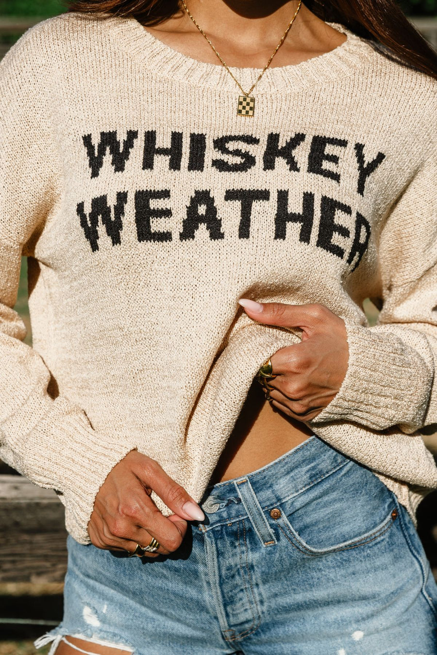 A person wearing the Panache Apparel Collection Tan Whiskey Weather Lightweight Sweater with “WHISKEY WEATHER” text and denim shorts is holding up part of the relaxed-fit sweater, revealing a necklace and rings on their fingers.