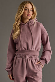 A woman with long blonde hair models the Dusty Purple Panache Cropped Hooded Sweatshirt by Panache Apparel Collection, paired with matching sweatpants, standing against a plain gray background and smiling slightly at the camera.
