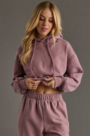 A woman with long blonde hair models the Panache Apparel Collection’s Dusty Purple Panache Cropped Hooded Sweatshirt, lifting the hem to reveal her midriff, paired with matching sweatpants against a plain gray background.