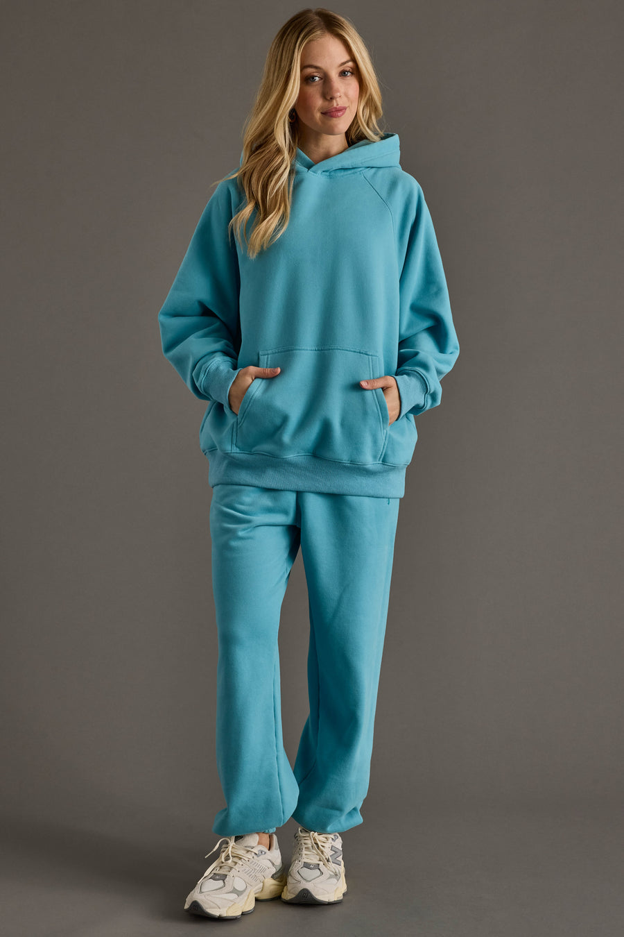 A woman with blonde hair smiles softly, standing against a gray background in Panache Apparel Collection's Bright Blue Panache Sweatpants and a matching hoodie, paired with white sneakers and hands in her front hoodie pocket.