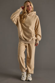 A person with long blonde hair wears a beige hoodie and Panache Apparel Collection's Tan Panache Sweatpants with an elastic waistband, paired with white sneakers, standing against a plain gray background.
