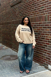 A woman with long, wavy hair stands against a brick wall wearing the Panache Apparel Collection Tan Whiskey Weather Lightweight Sweater, loose blue jeans, and black sandals, looking slightly to the side.