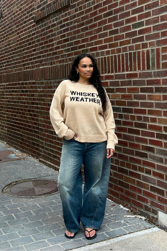 A woman with long, wavy hair stands against a brick wall wearing the Panache Apparel Collection Tan Whiskey Weather Lightweight Sweater, loose blue jeans, and black sandals, looking slightly to the side.