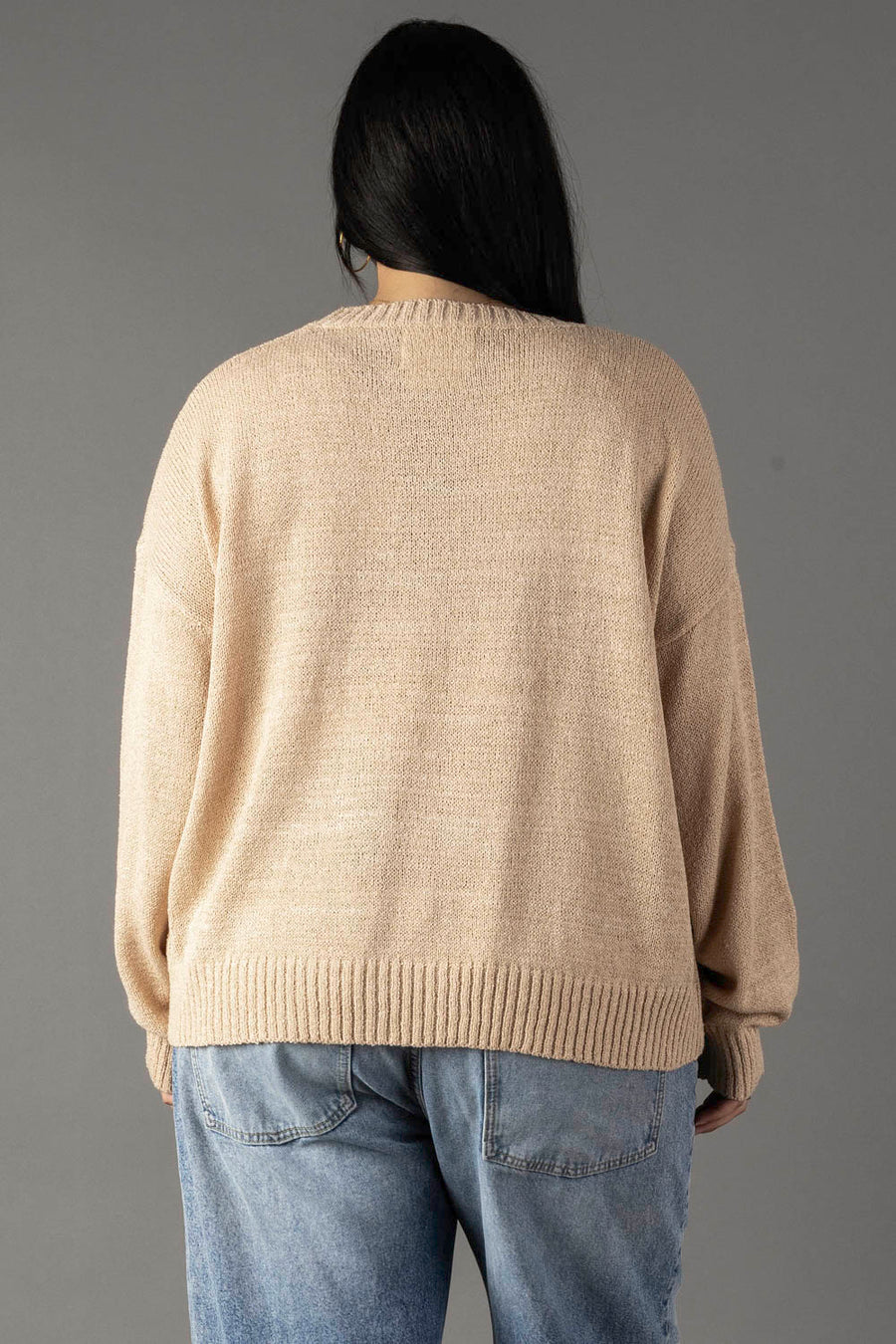 A person with long dark hair is seen from behind, wearing the Panache Apparel Collection’s Tan & Ivory Coffee Weather Lightweight Sweater and light blue jeans, standing against a plain gray background.