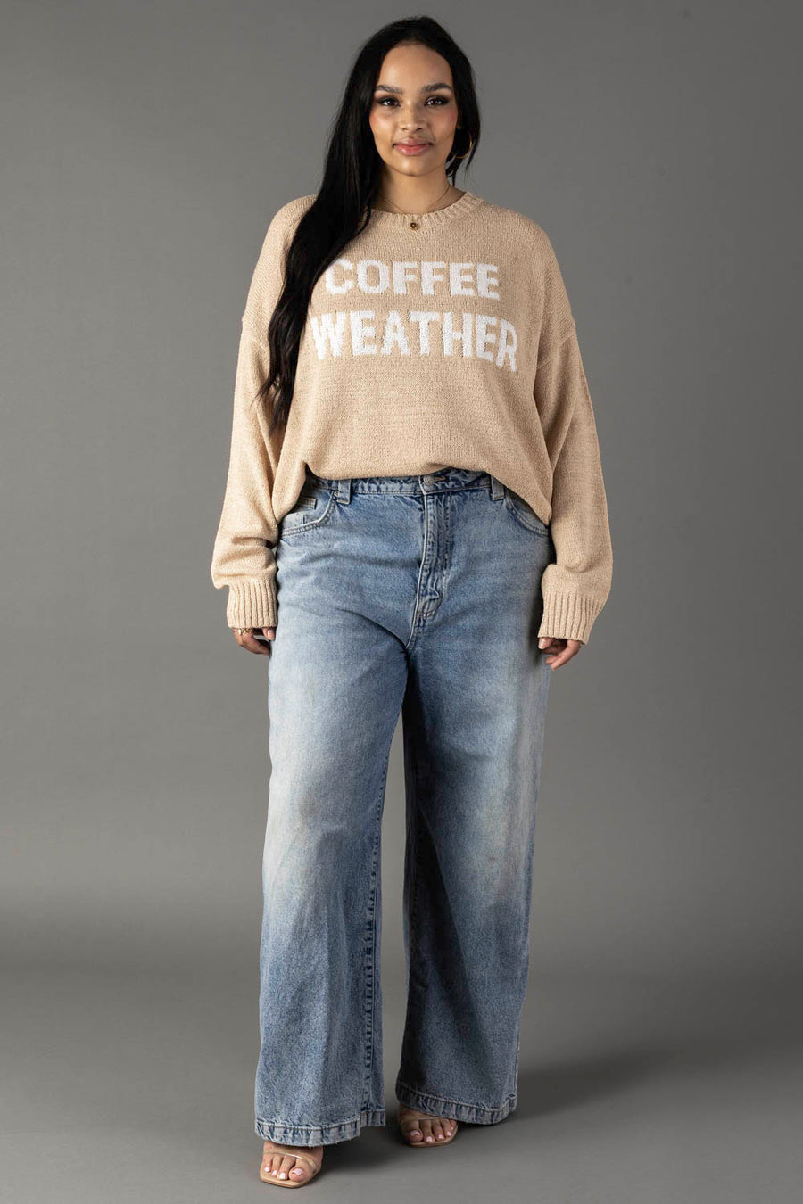 A woman with long dark hair smiles softly against a gray background, wearing Panache Apparel Collection's Tan & Ivory Coffee Weather Lightweight Sweater and loose blue jeans.