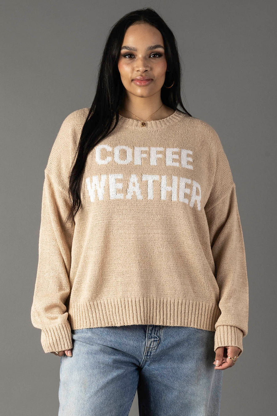 A woman with long dark hair wears the Panache Apparel Collection’s Tan & Ivory Coffee Weather Lightweight Sweater—featuring white lettering—paired with light blue jeans, standing before a plain gray background.