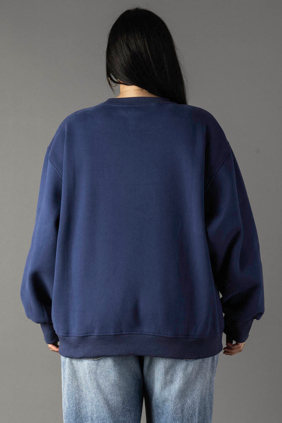 A person with long dark hair, seen from behind, wears the Panache Apparel Collection's Drink At The Cabin Sweatshirt in navy blue with light blue jeans, standing against a plain gray background.