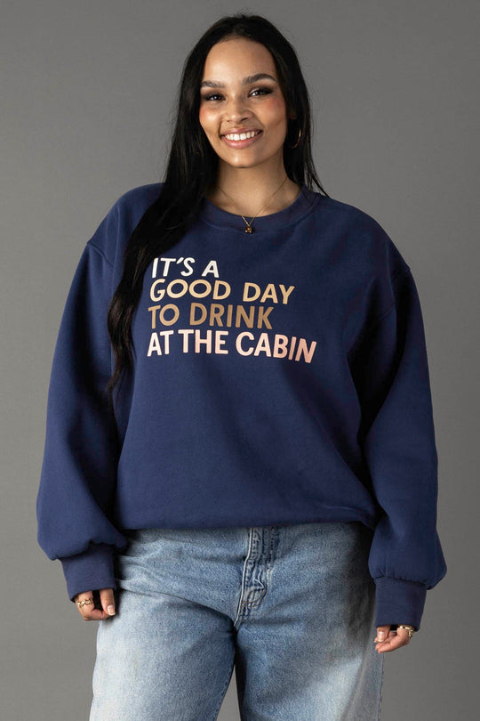 A woman with long dark hair smiles in a Panache Apparel Collection Drink At The Cabin Sweatshirt, an oversized navy top with "It’s a good day to drink at the cabin" in tan and beige text. She pairs it with light blue jeans against a plain gray background.
