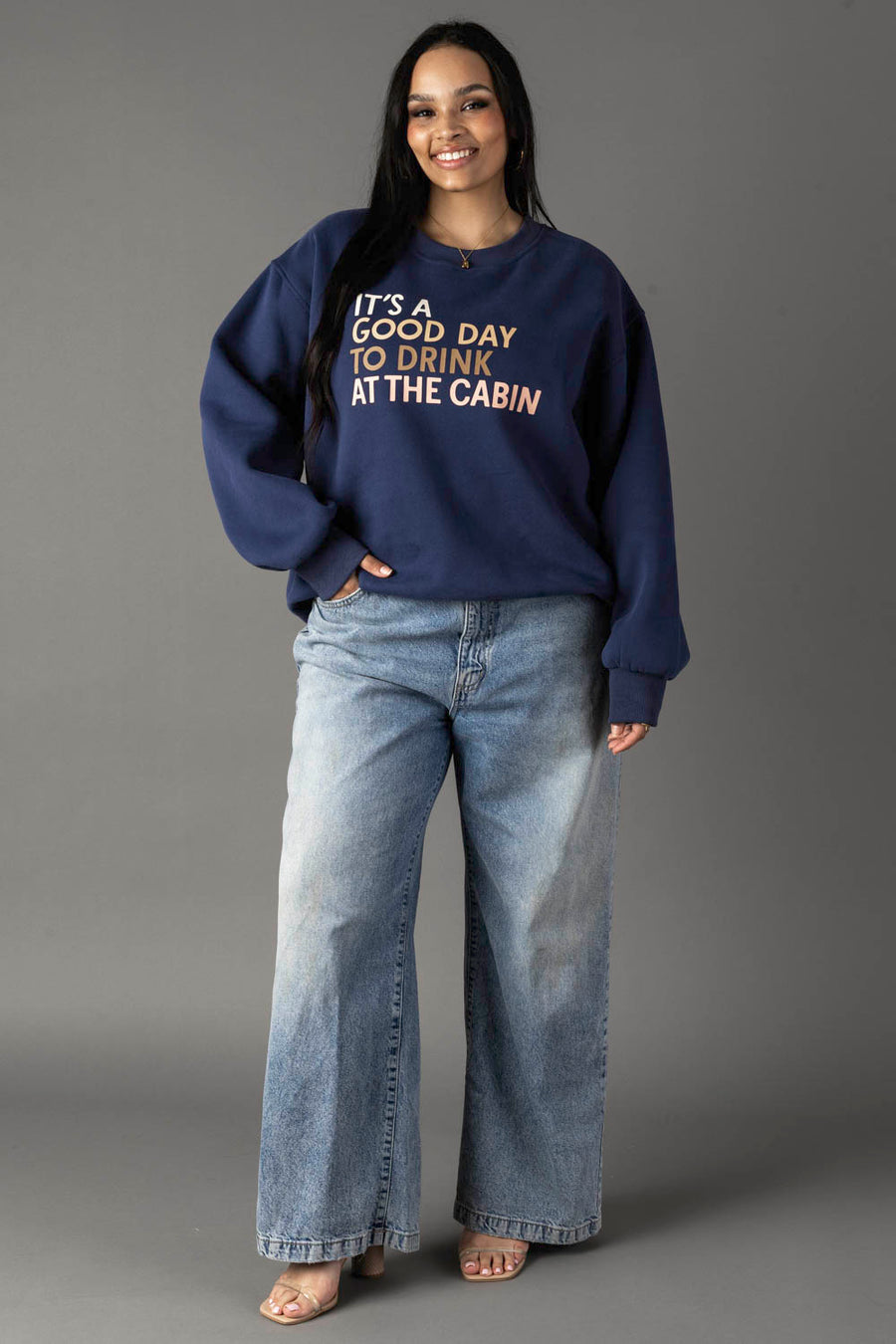 A woman smiles in front of a gray background, wearing the Panache Apparel Collection "Drink At The Cabin Sweatshirt," paired with blue jeans and beige sandals.