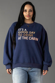 A woman with long black hair wears the Panache Apparel Collection Drink At The Cabin Sweatshirt in navy blue, featuring “ITS A GOOD DAY TO DRINK AT THE CABIN,” paired with light blue jeans against a plain gray background.