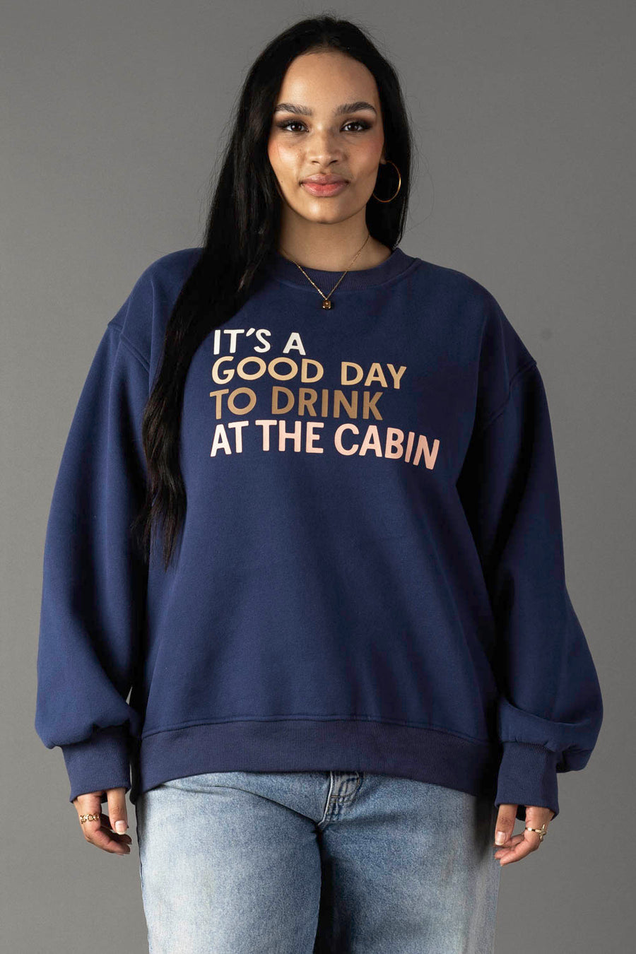 A woman with long black hair wears the Panache Apparel Collection Drink At The Cabin Sweatshirt in navy blue, featuring “ITS A GOOD DAY TO DRINK AT THE CABIN,” paired with light blue jeans against a plain gray background.