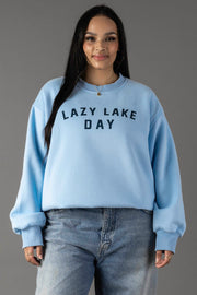 A woman smiles in a light blue Lazy Lake Day Sweatshirt by Panache Apparel Collection, paired with matching jeans against a gray background.