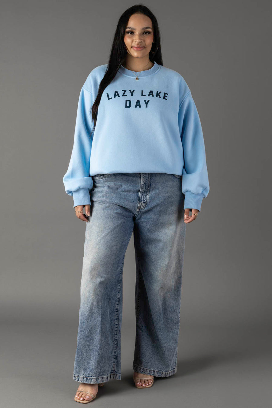 A woman stands against a gray background, smiling with her hands by her sides, wearing an oversized Panache Apparel Collection Lazy Lake Day Sweatshirt and loose-fitting blue jeans.