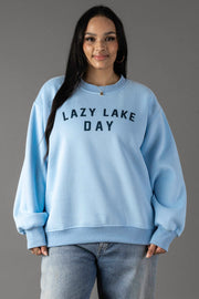 A woman with long dark hair stands against a gray background, smiling gently. She wears the Panache Apparel Collection’s Lazy Lake Day Sweatshirt in light blue with matching jeans, giving her a relaxed and effortlessly stylish look.