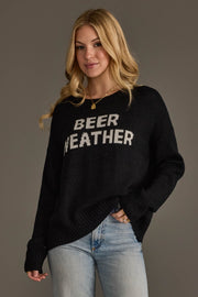 A woman with long blonde hair wears the Panache Apparel Collection Beer Weather Lightweight Sweater in black, paired with light blue jeans and gold jewelry, standing against a plain gray background.