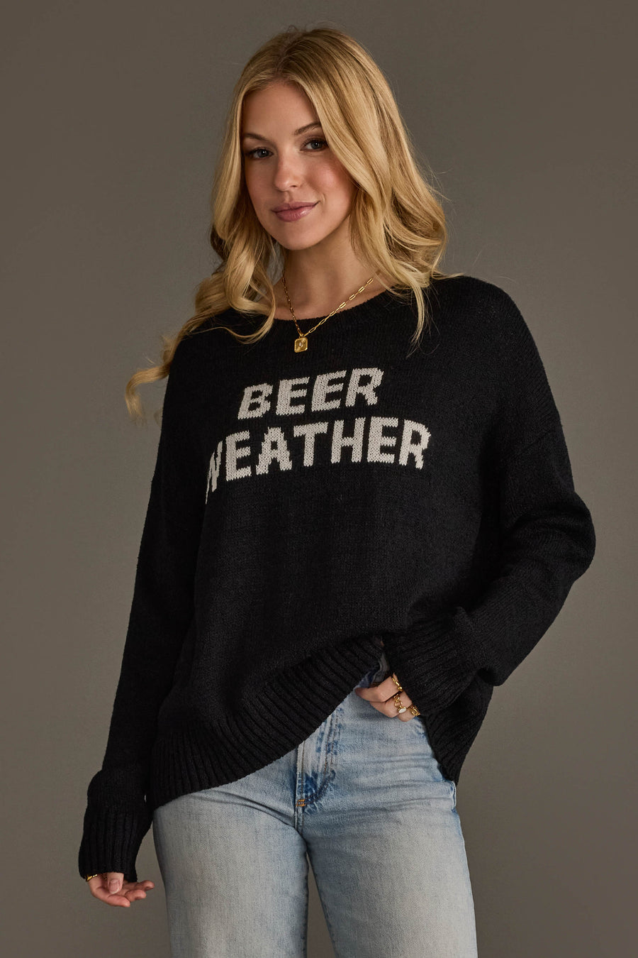 A woman with long blonde hair wears the Panache Apparel Collection Beer Weather Lightweight Sweater in black, paired with light blue jeans and gold jewelry, standing against a plain gray background.