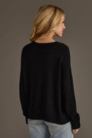 A woman with blonde hair models the Panache Apparel Collection's Beer Weather Lightweight Sweater in black with light blue jeans, standing facing away and looking to the side against a plain dark background.