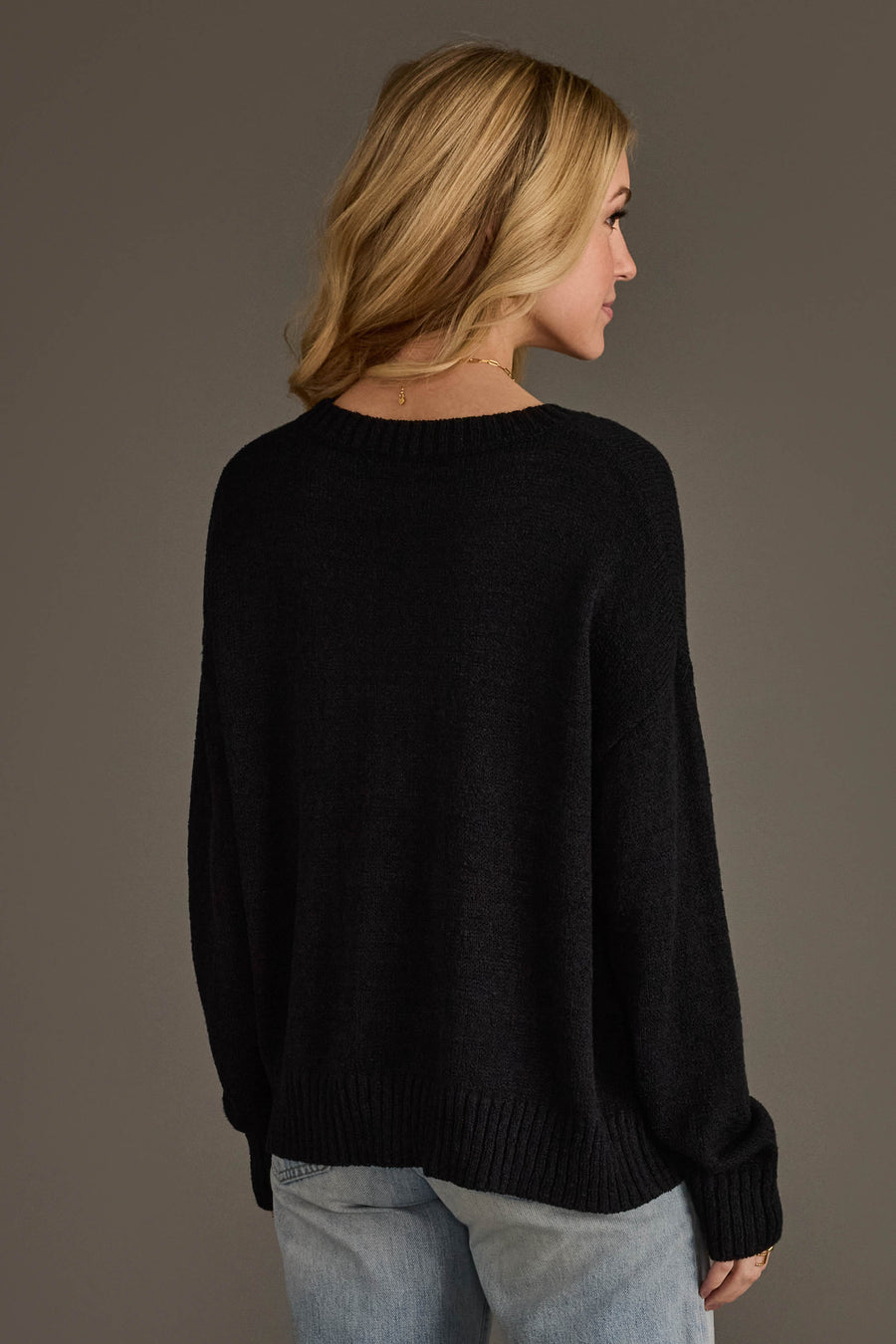 A woman with blonde hair models the Panache Apparel Collection's Beer Weather Lightweight Sweater in black with light blue jeans, standing facing away and looking to the side against a plain dark background.