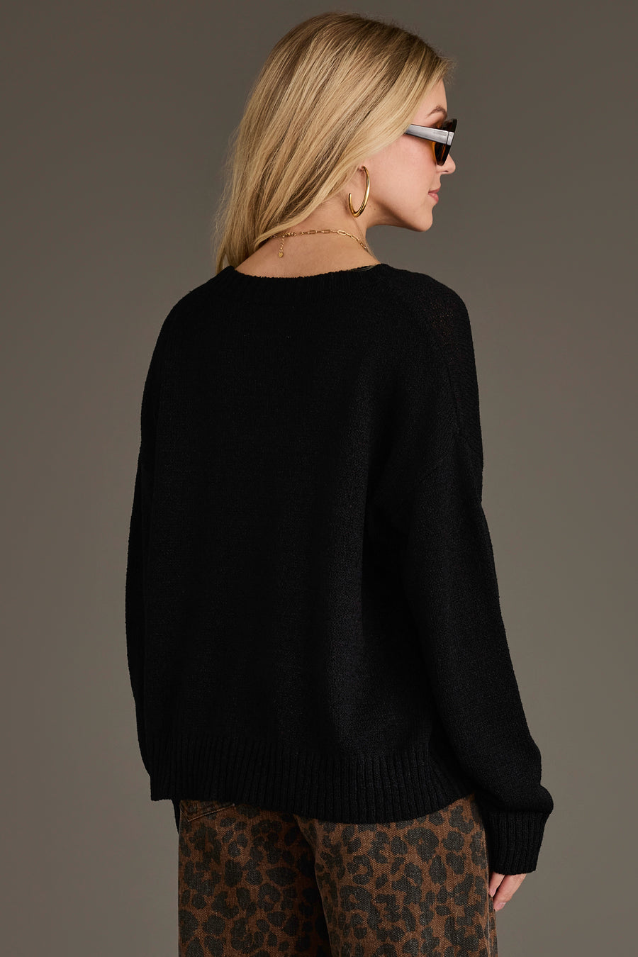 A woman with long blonde hair, sunglasses, and gold hoop earrings stands facing away from the camera against a plain background, wearing the Panache Apparel Collection’s Beer Weather Lightweight Sweater in black and brown leopard-print pants.
