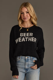 A woman with long blonde hair wears the Beer Weather Lightweight Sweater by Panache Apparel Collection, featuring white lettering, paired with light blue jeans. She stands against a plain gray background and smiles slightly.