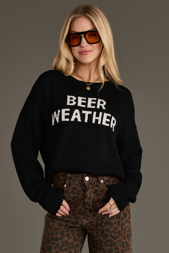 A woman wearing the Panache Apparel Collection’s Beer Weather Lightweight Sweater in black and leopard print pants stands with one hand in her pocket, sunglasses on, against a plain background.