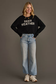 A smiling woman stands with her hands in her pockets, wearing Panache Apparel Collection's Beer Weather Lightweight Sweater in black, light blue high-waisted jeans, and white sneakers against a plain gray background.